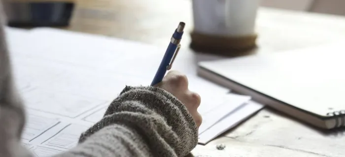 A person writing with a pen at a wooden table with a mug of coffee nearby