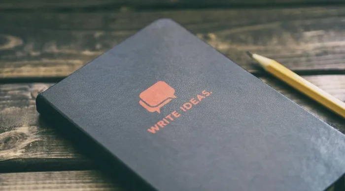 A dark notebook reading Write Ideas resting on a wooden table with a pencil