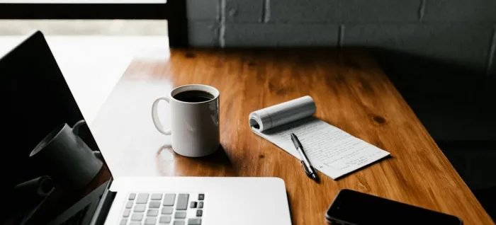 A laptop, coffee mug, and notepad with a pen on a wooden desk
